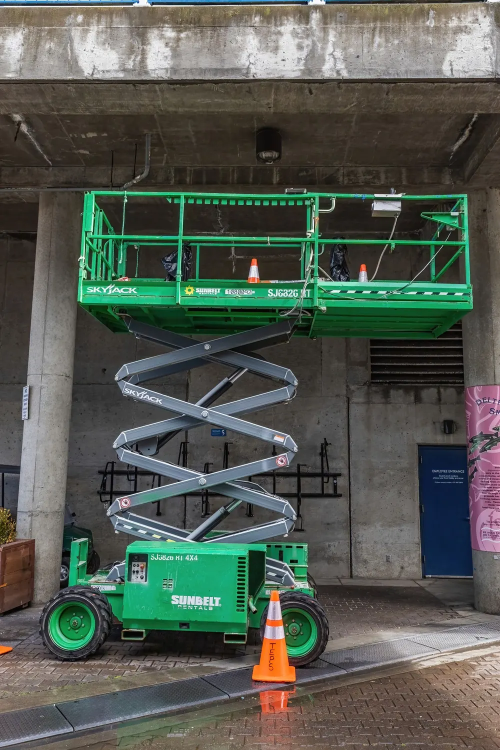 Green scissor lift parked beside a building with safety cones