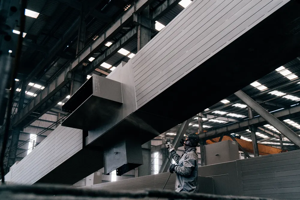 Worker in protective suit spray coating large metal beams in a warehouse